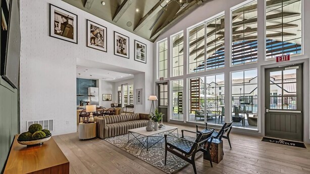 A spacious living area with a large window, a sofa, a dining table, and a kitchen in the background.