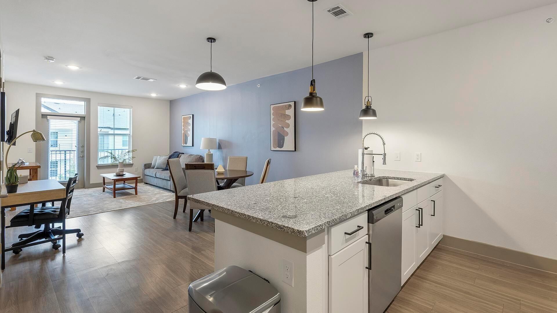 A modern kitchen with granite countertops, a dishwasher, and a sink. There is a dining area with a table and chairs, and a living room with a couch and a coffee table.