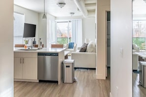 A modern kitchen with a dishwasher, a small trash bin, and a dining area with a sofa. - Landing Apartments Salt Lake City (Salt Lake City)