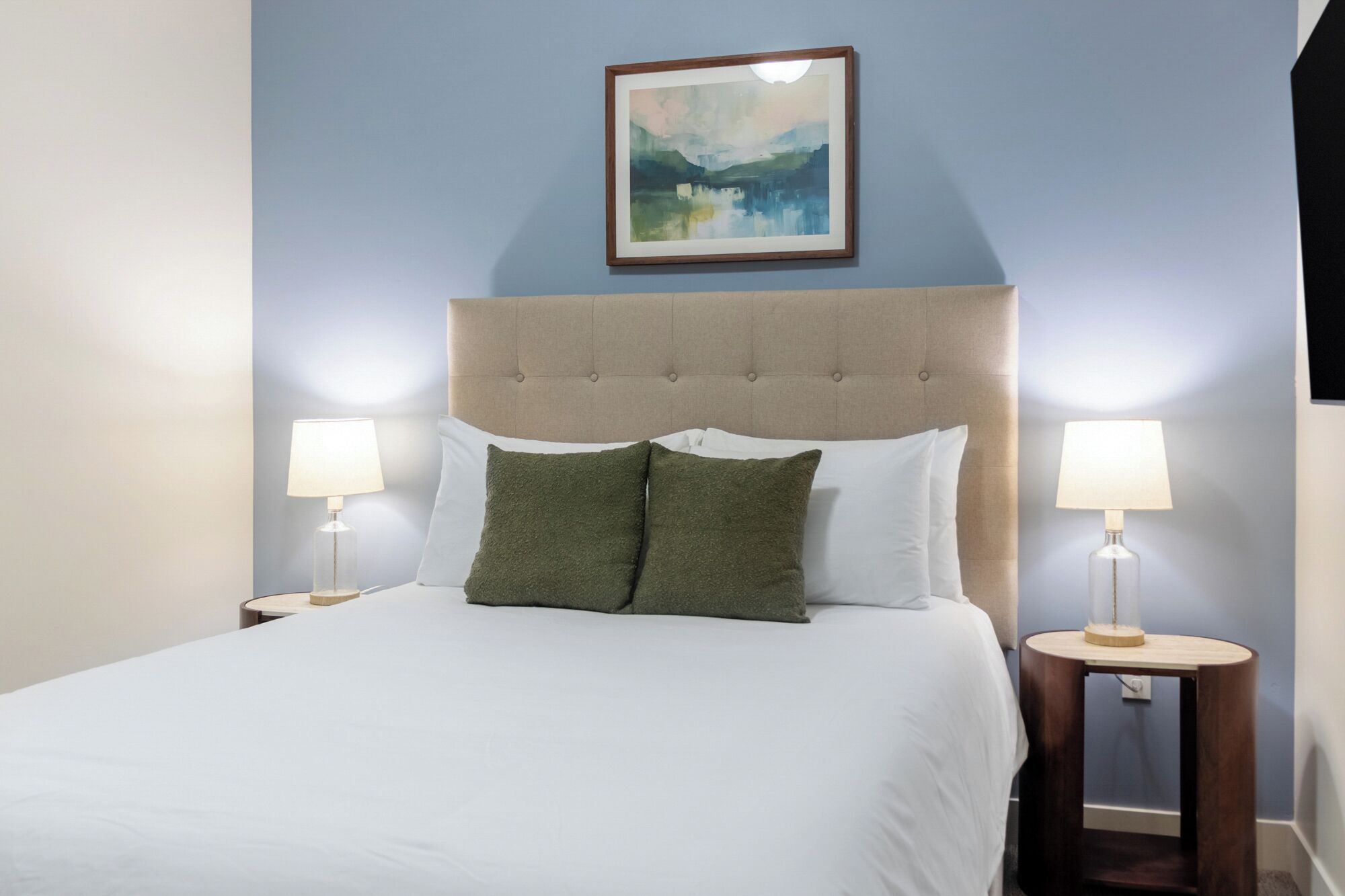 A neatly made bed with a tufted headboard, two bedside lamps, and a framed painting above.