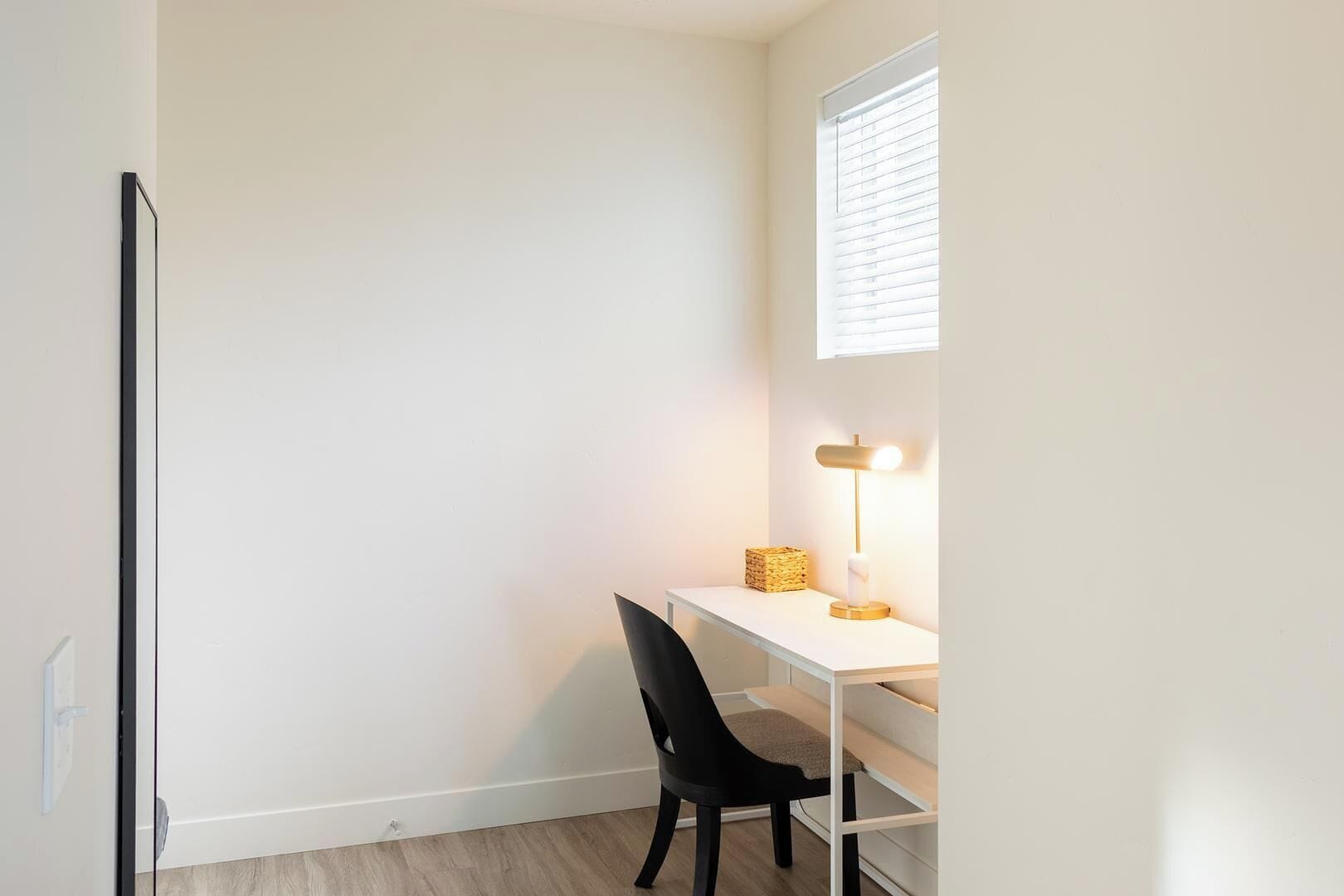 A small, bright room with a white desk, a black chair, a lamp, and a window with blinds.