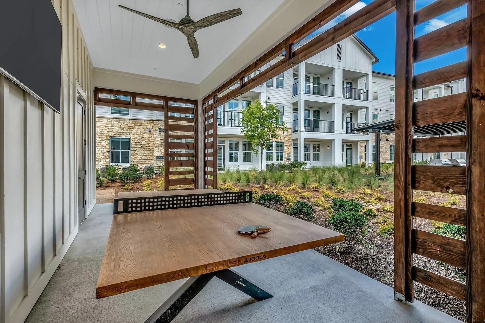 A modern outdoor seating area with a wooden table and chairs, a ceiling fan, and a view of a multi-story building with balconies.