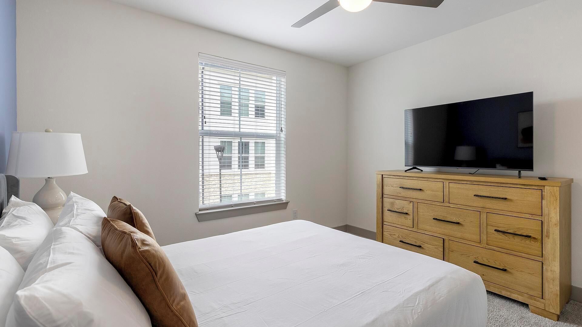 Een slaapkamer met een bed, een dressoir, een televisie en een raam met jaloezieën.