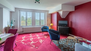 Ein Wohnzimmer mit rotem Teppich, einem Essbereich mit Tisch und Stühlen, einem Fernseher und einem Fenster mit Blick auf Bäume.