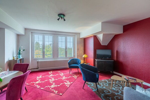 A living room with a red carpet, a dining area with a table and chairs, a TV, and a window with a view of trees.