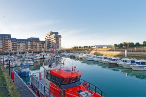 A marina with multiple boats docked, a prominent red boat in the foreground, and a multi-story building in the background. - A Joaninha - Vue sur le Port (Courseulles-sur-Mer)
