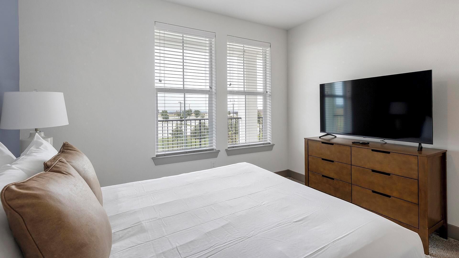 Een slaapkamer met een bed, een tv op een houten dressoir en twee ramen met jaloezieën.