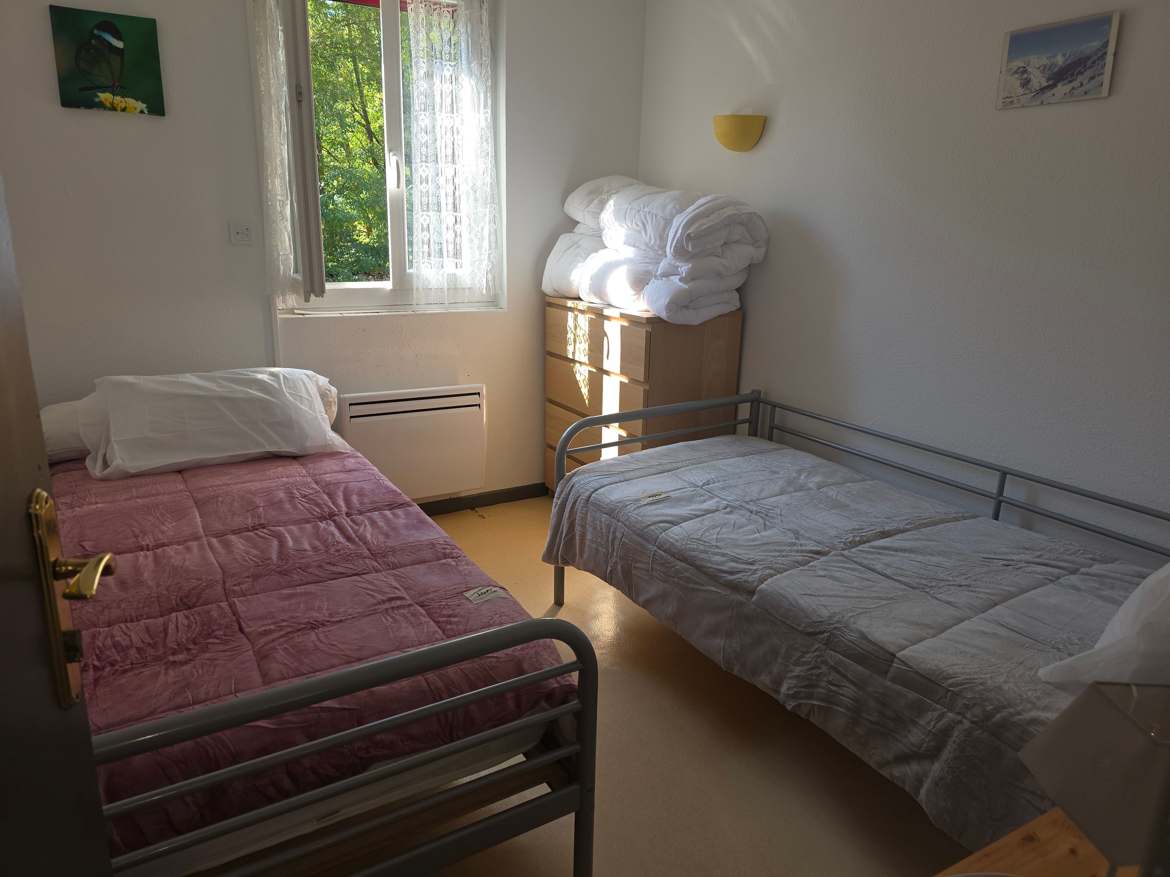 Une chambre de dortoir avec deux lits, une fenêtre avec des rideaux, un radiateur fixé au mur et un petit tableau.