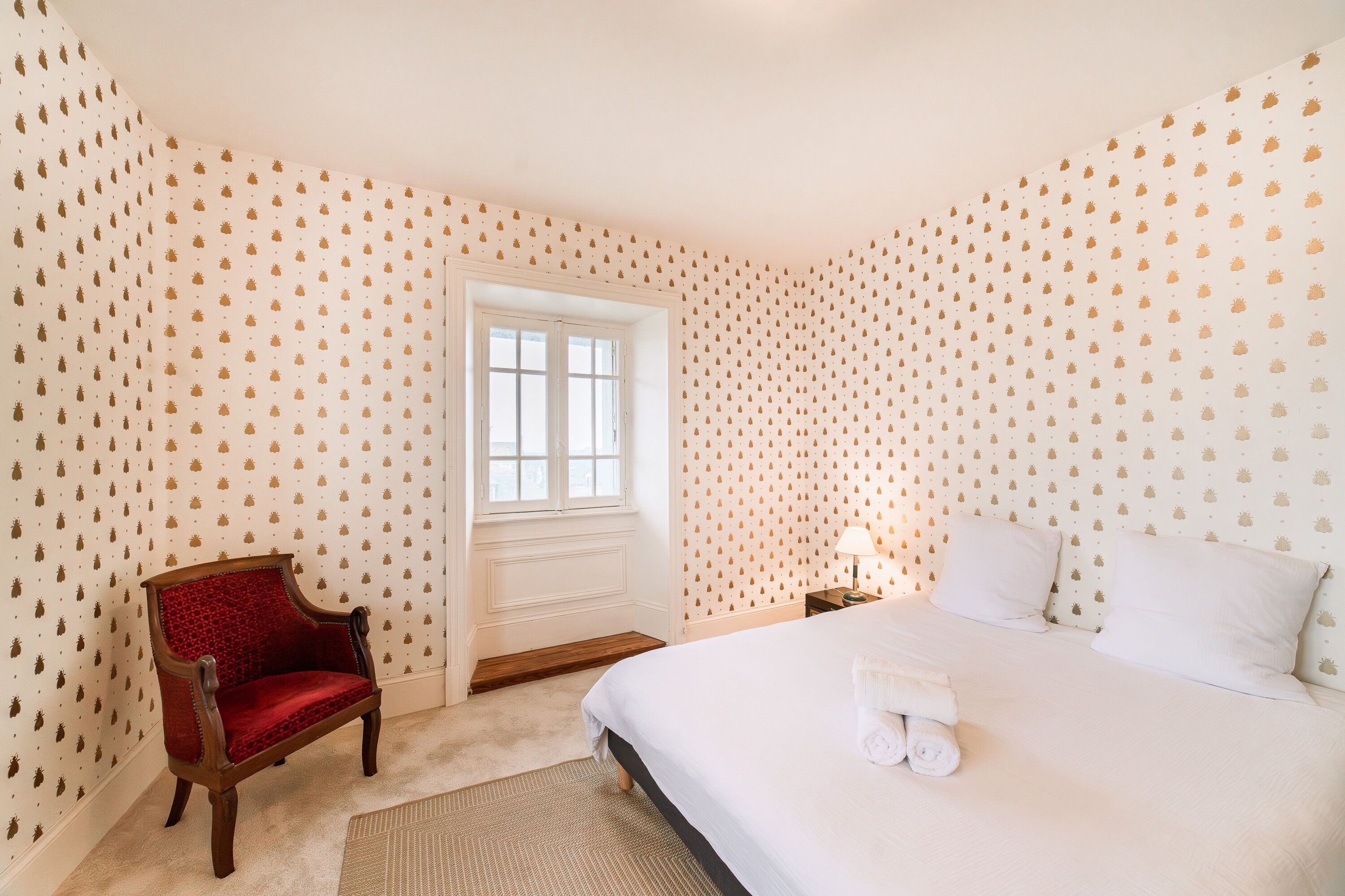 Un dormitorio con cama, una silla, un ventanal y papel tapiz estampado.