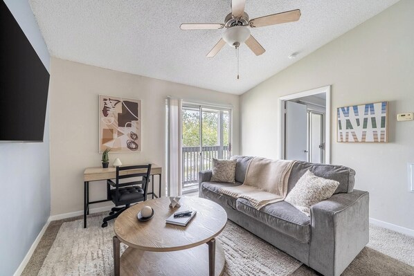 A living room with a gray sofa, a round wooden coffee table, a desk with a chair, and a ceiling fan. - Landing | Spectacular 1BD, Gym, Tennis Court (Palm Bay)