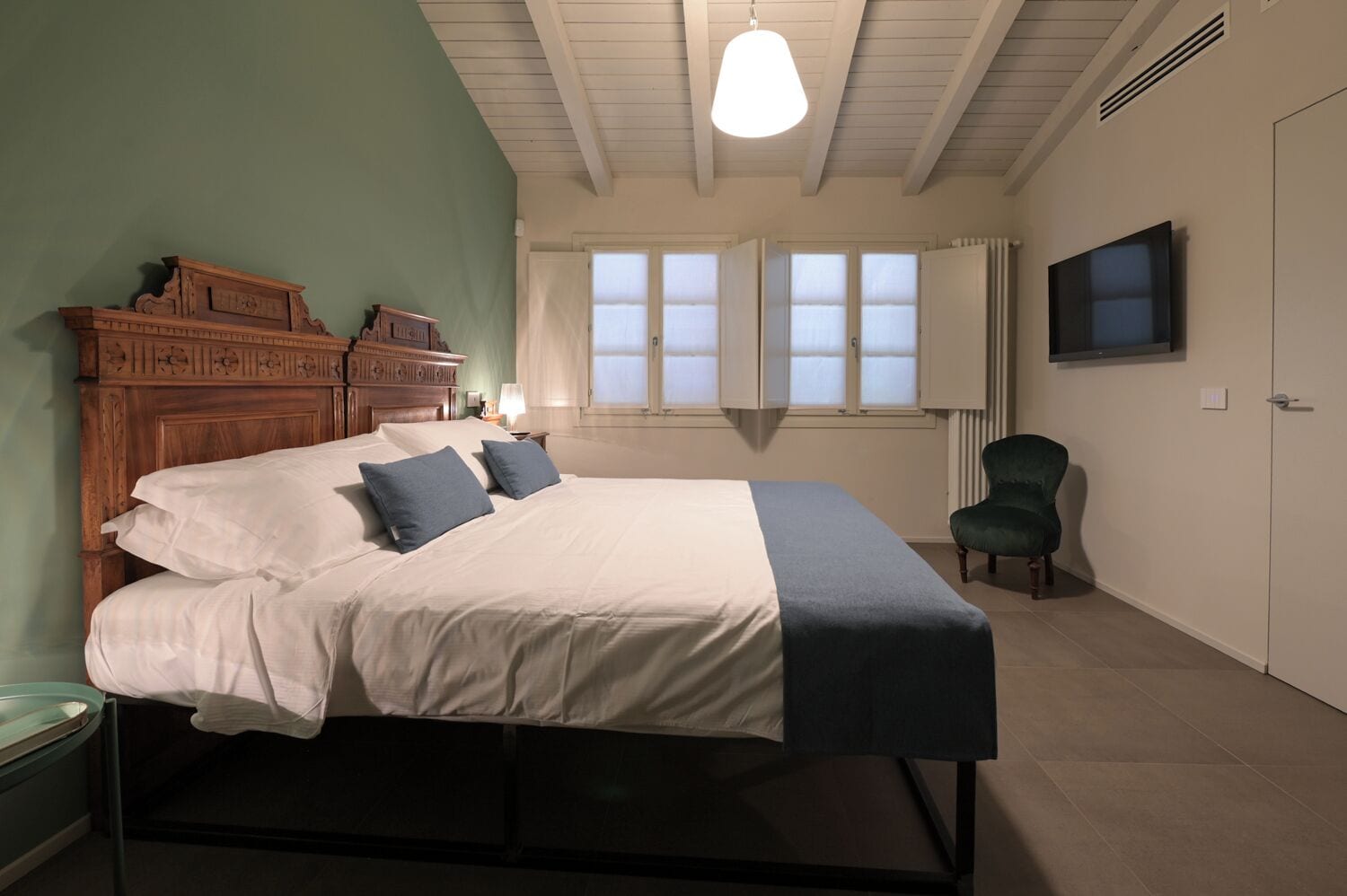 A bedroom with a bed, a wooden headboard, a green armchair, a TV, and a window with curtains.