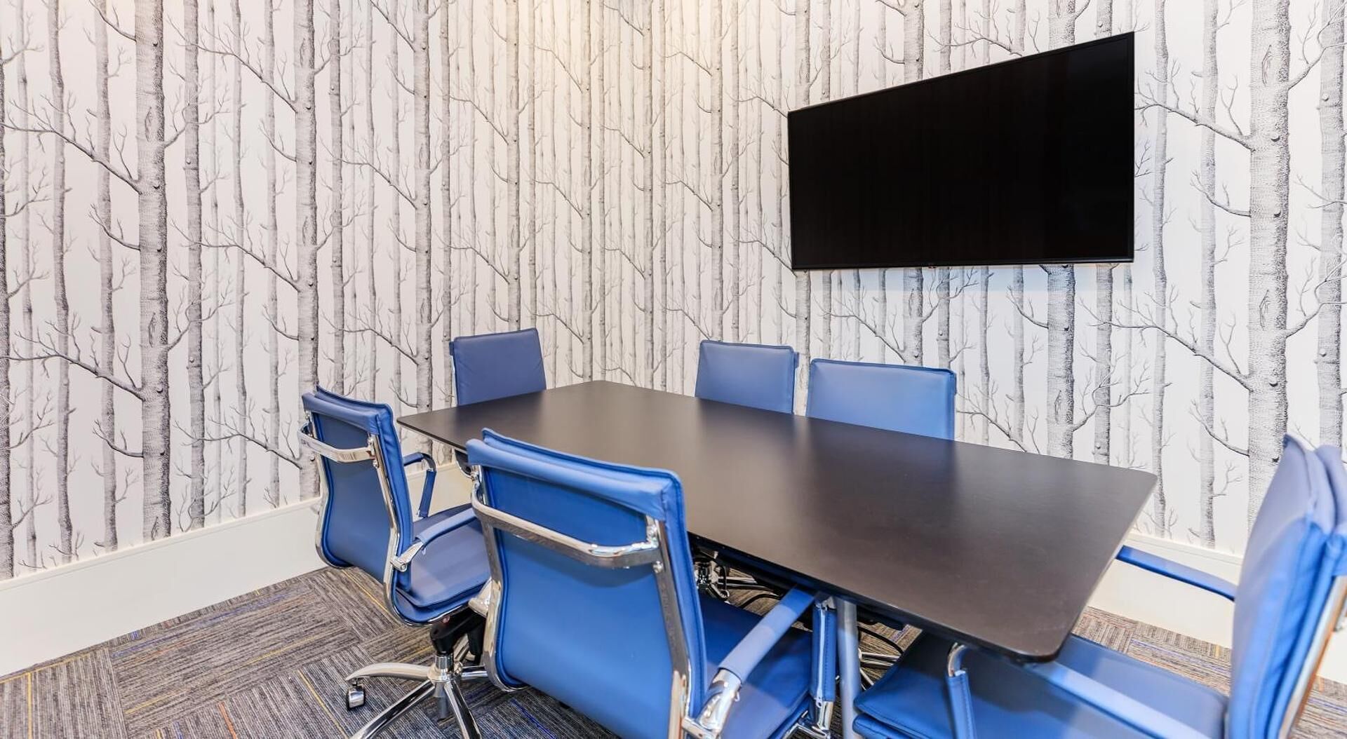 Ein Konferenzraum mit dunklem Tisch, blauen Stühlen und einem an der Wand befestigten Flachbildfernseher.