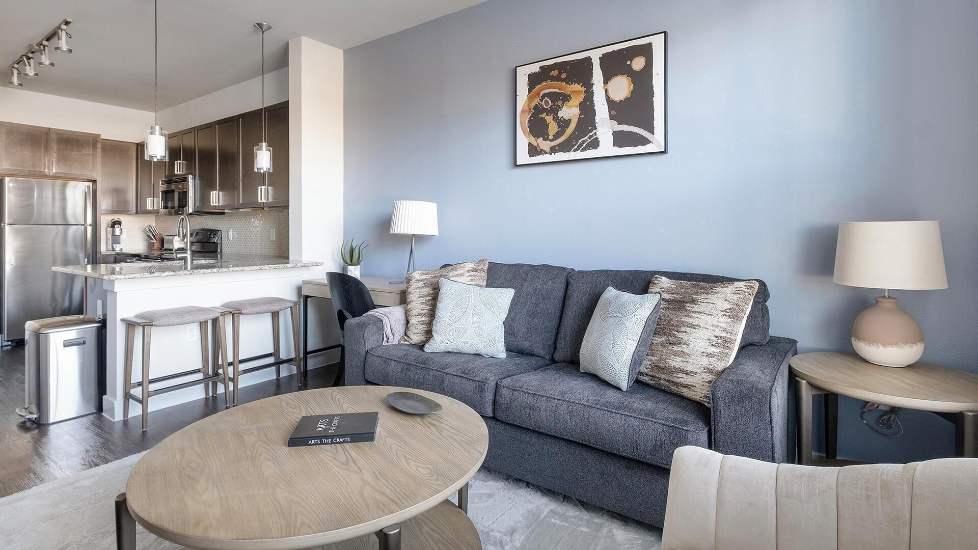 A modern living room with a grey sofa, a round wooden coffee table, and a kitchen area with stainless steel appliances.