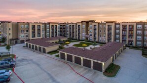 A multi-story apartment complex with a central courtyard, parking area, and a row of single-story buildings with attached garages. - Landing | Spectacular 1BD, Pool, Gym (Houston)
