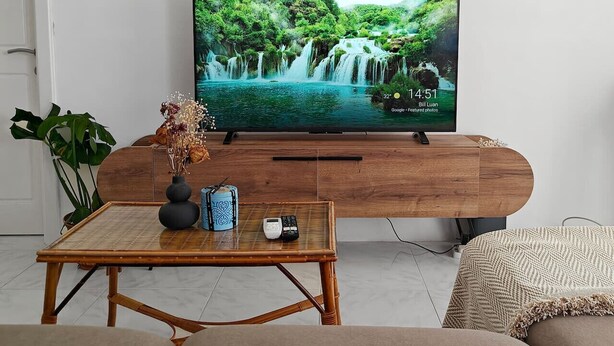 Ein Wohnzimmer mit Fernseher, der eine Wasserfallszene zeigt, einem Holztisch mit Vase und Fernbedienung sowie einem Sofa.