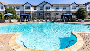 A swimming pool with lounge chairs and umbrellas, surrounded by a building with multiple windows and stone accents. - Landing | Beautiful 1BD, Clubhouse, Pool (Greenville)