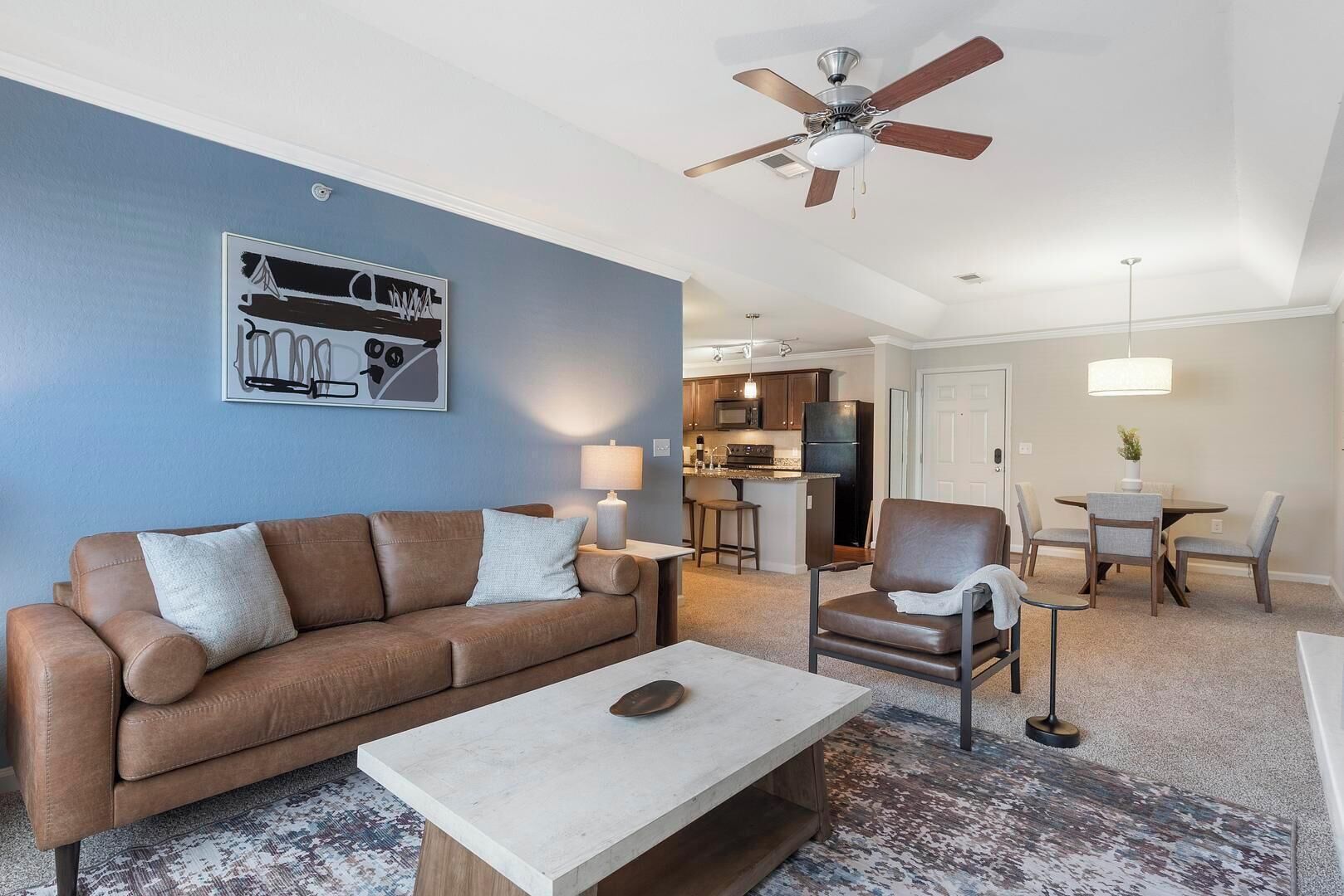 A living room with a brown leather sofa, a coffee table, and a ceiling fan. There is a kitchen in the background with a dining area.