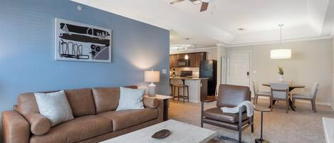 A living room with a brown leather sofa, a coffee table, and a ceiling fan. There is a kitchen in the background with a dining area.