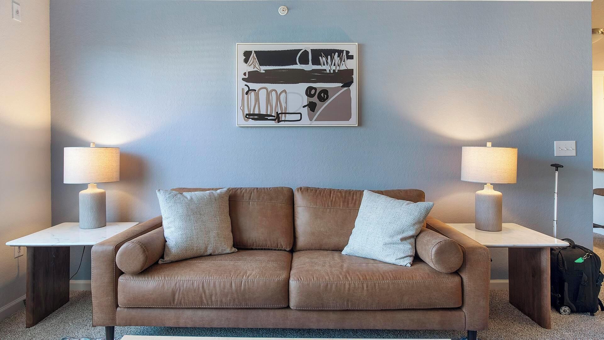 Una sala de estar moderna con un sofá de cuero marrón, dos mesitas de noche con lámparas y una obra de arte abstracto en la pared.