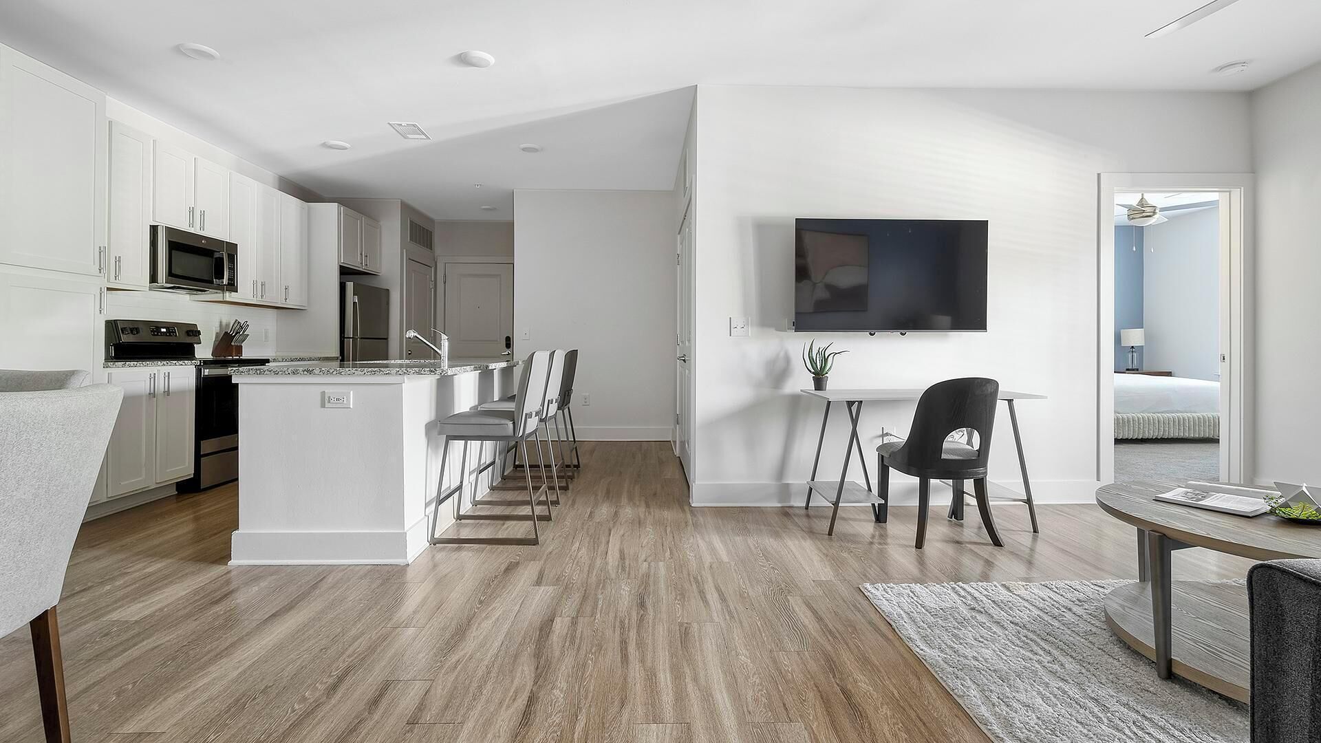 Una cocina moderna con armarios blancos, un microondas y una estufa. Hay un comedor con una mesa y sillas, y un televisor montado en la pared.