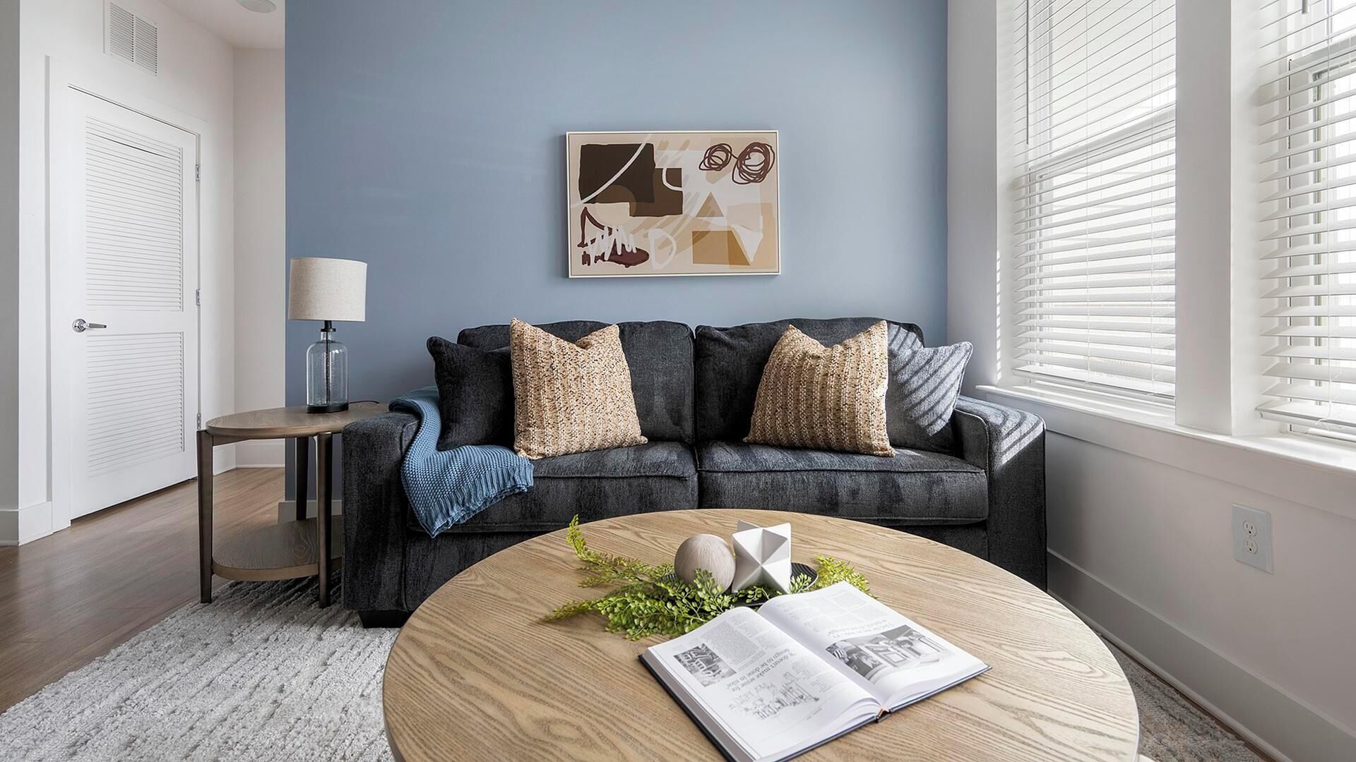 A modern living room with a dark grey sofa, a round wooden coffee table, and a framed abstract artwork on the wall.