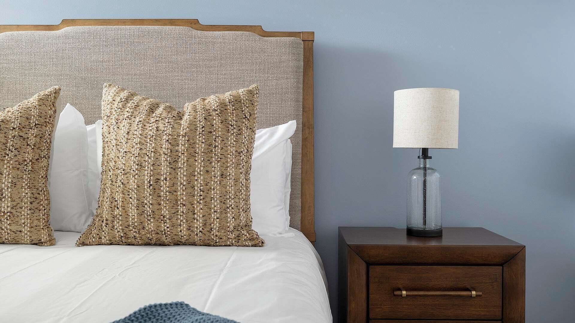 A bed with a woven headboard, three patterned pillows, a white comforter, a blue throw, and a bedside table with a lamp.