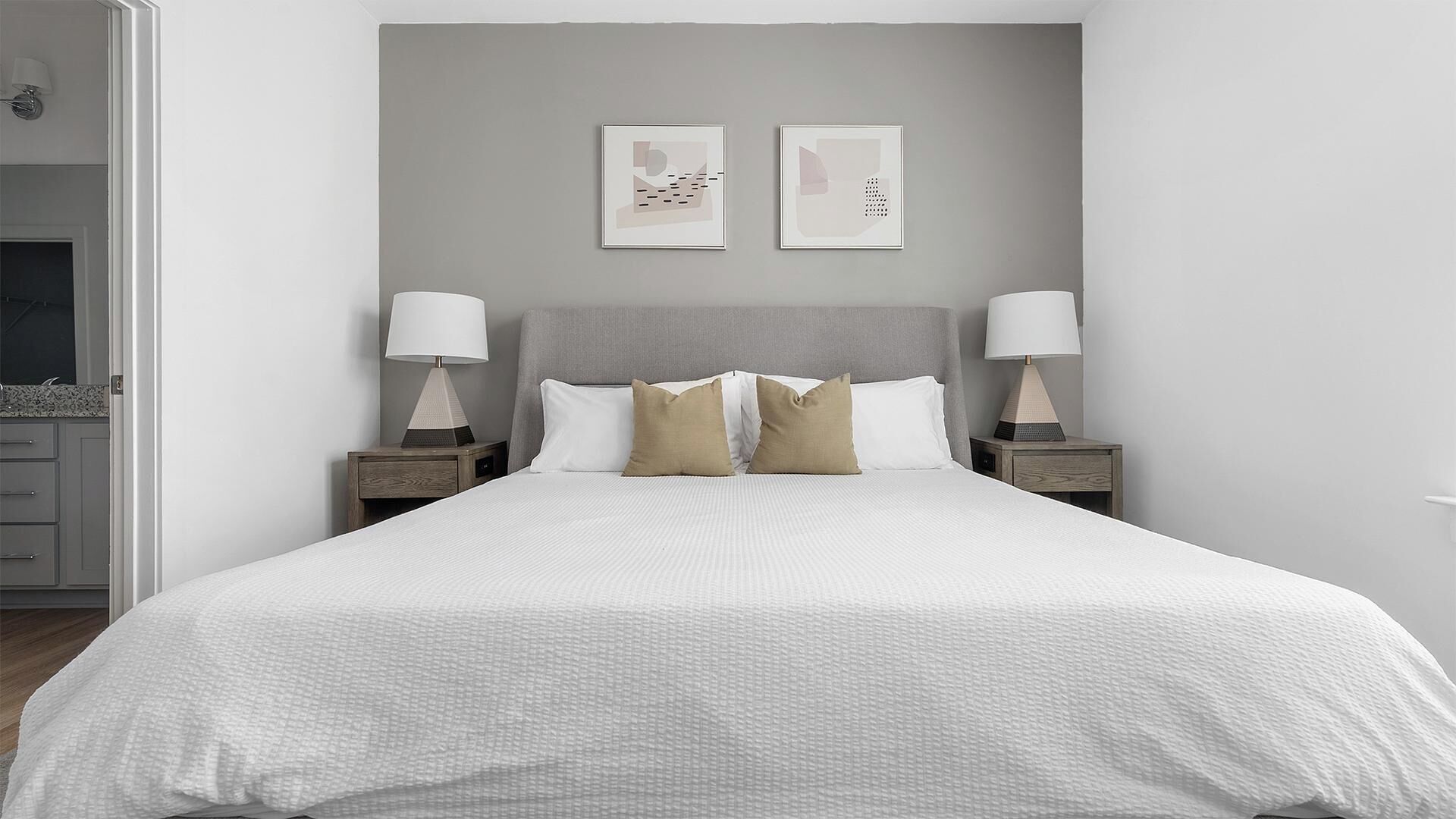 A neatly made bed with a grey headboard, two bedside tables with lamps, and two framed artworks on the wall.