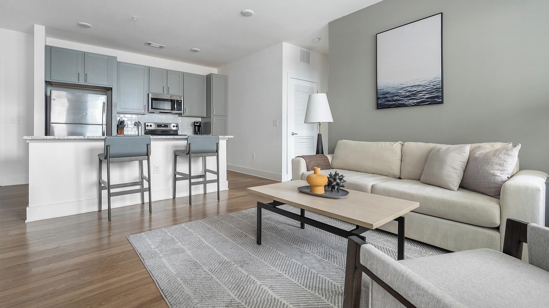 A modern kitchen with grey cabinets and stainless steel appliances, a dining area with a table and chairs, and a living room with a sofa and a coffee table.