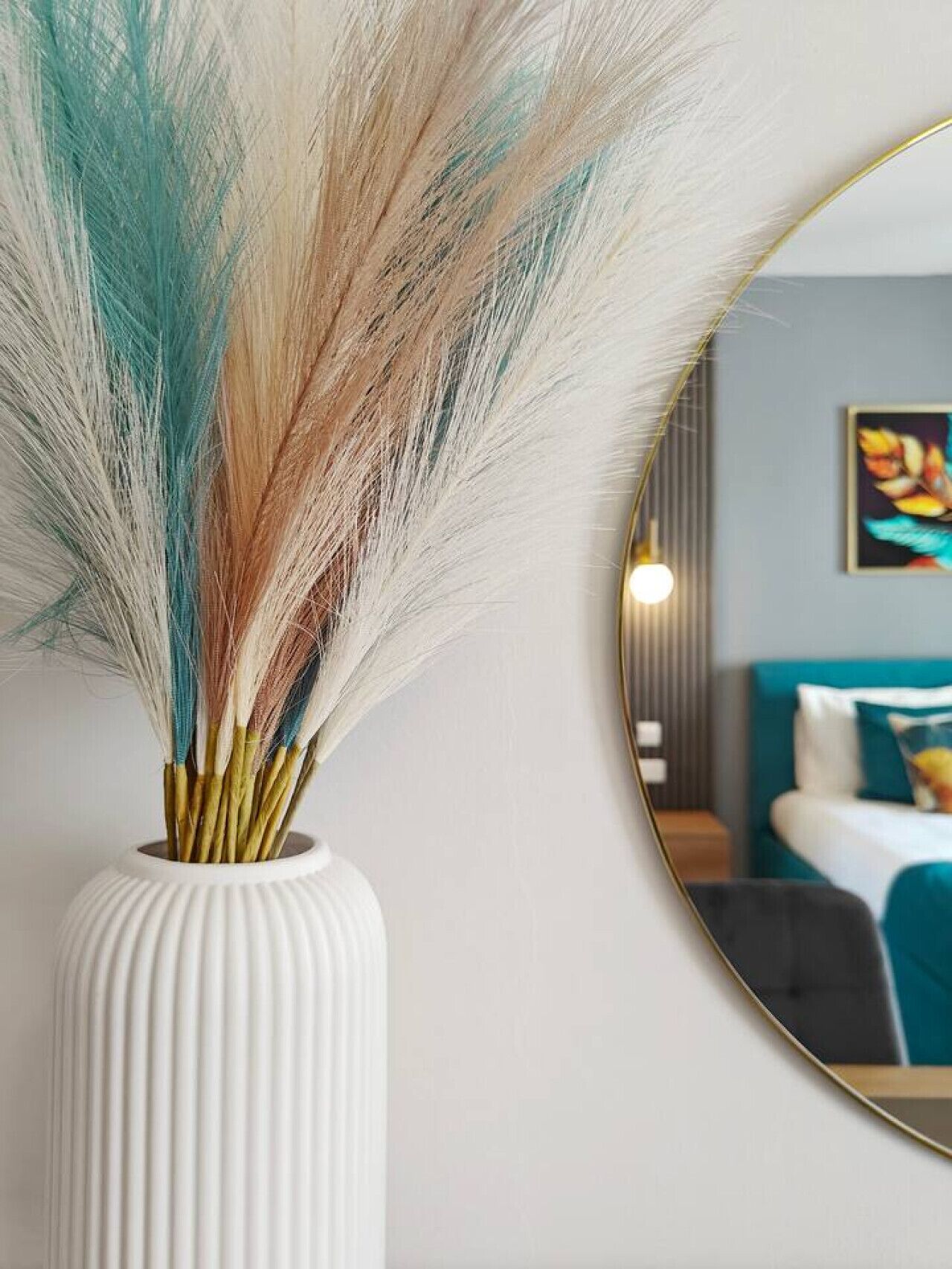 A white vase with a ribbed texture containing colorful feathers, placed next to a round mirror reflecting a room with a bed and a painting on the wall.