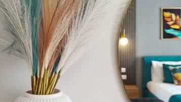 A white vase with a ribbed texture containing colorful feathers, placed next to a round mirror reflecting a room with a bed and a painting on the wall.