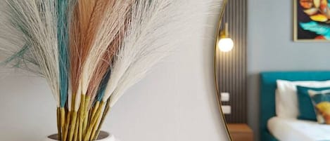 Un jarrón blanco con textura acanalada que contiene plumas de colores, colocado junto a un espejo redondo que refleja una habitación con una cama y un cuadro en la pared.