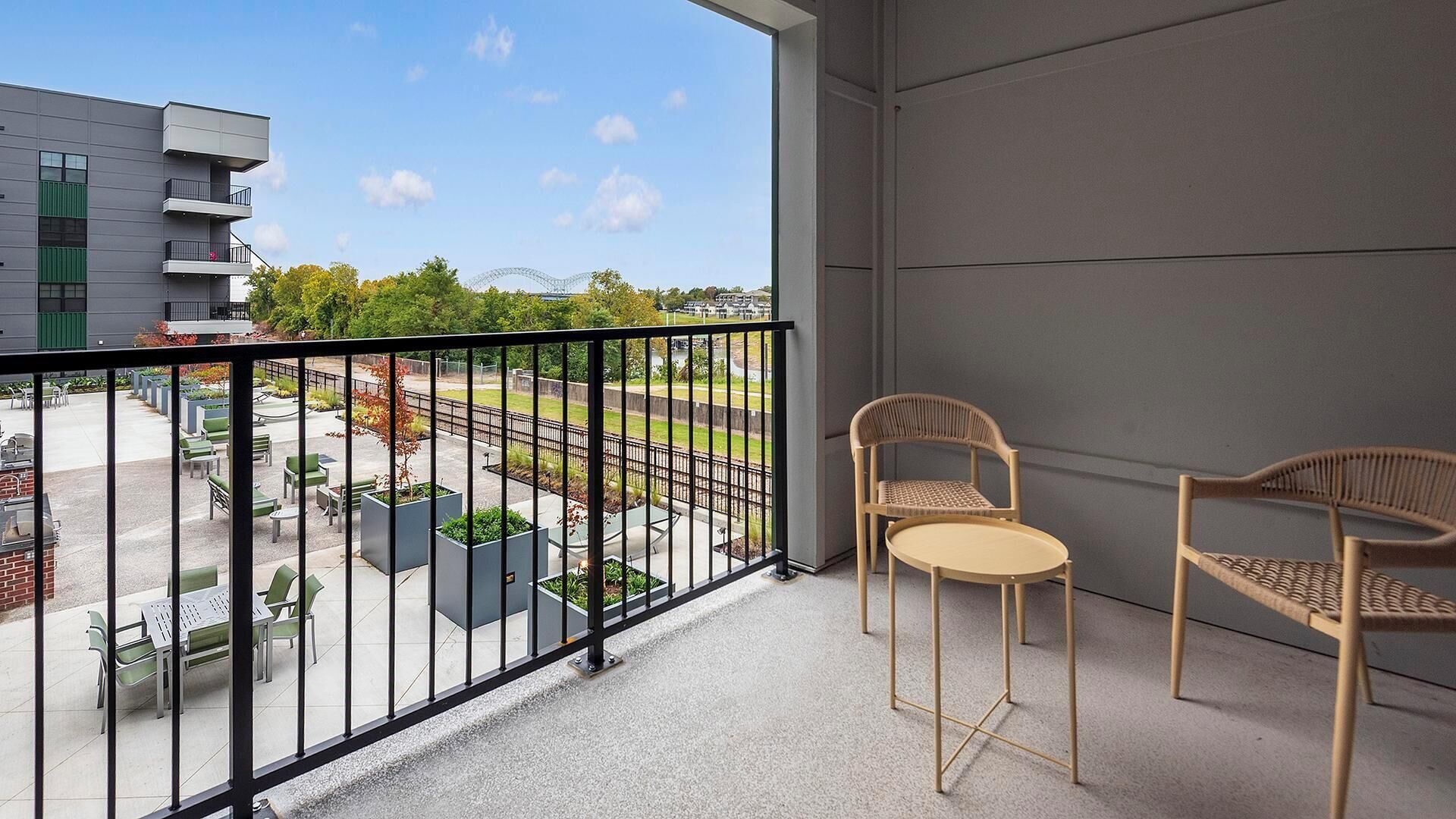 A balcony with two wicker chairs, a view of a building with balconies, and a fenced outdoor area with seating and planters.