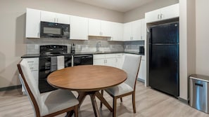 Eine moderne Küche mit weißen Schränken, einem schwarzen Kühlschrank, einer Mikrowelle, einem Backofen und einem Geschirrspüler. Ein runder Holztisch wird von zwei Stühlen begleitet.