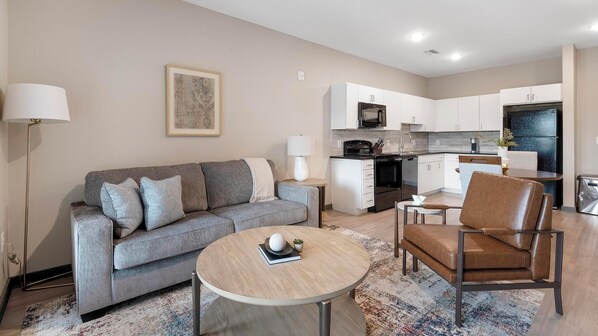 Um apartamento moderno com sofá cinzento, mesa redonda de madeira, poltrona de couro marrom, cozinha com armários brancos e um pequeno espaço para refeições.