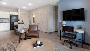 A modern living room with a round coffee table, a brown leather chair, a desk with a chair, a flat-screen TV mounted on the wall, and a kitchen area with cabinets and a refrigerator.