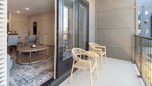 A balcony with a round table and two wicker chairs, a door with a window, and a view of a building. - Landing | Incredible 1BD, Rooftop Lounge, Pool (Memphis)