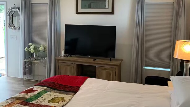 A hotel room with a bed, a television on a wooden stand, a framed picture on the wall, and a window with curtains. - Fully Furnished Studio Directly Across from TMH (Tallahassee)