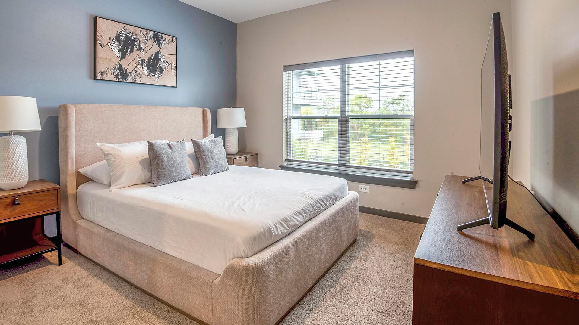 A bedroom with a bed, bedside tables, a TV, and a window with blinds.