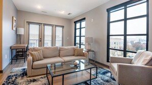 A modern living room with a beige sofa, a glass coffee table, and a view of the city through large windows. - Landing | Amazing 1BD, Rooftop Lounge, Yoga Studio (Memphis)