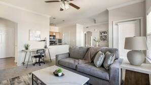 A modern living room with a grey sofa, a coffee table, and a lamp. There is a kitchen in the background with wooden cabinets and a ceiling fan. - Landing | Spectacular 1BD, Clubhouse, Pool (Grand Prairie)
