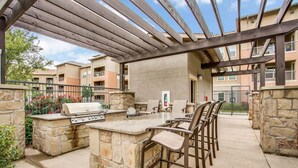 Eine Terrasse im Freien mit Grillplatz aus Stein, Sitzgelegenheiten und einer Pergola.