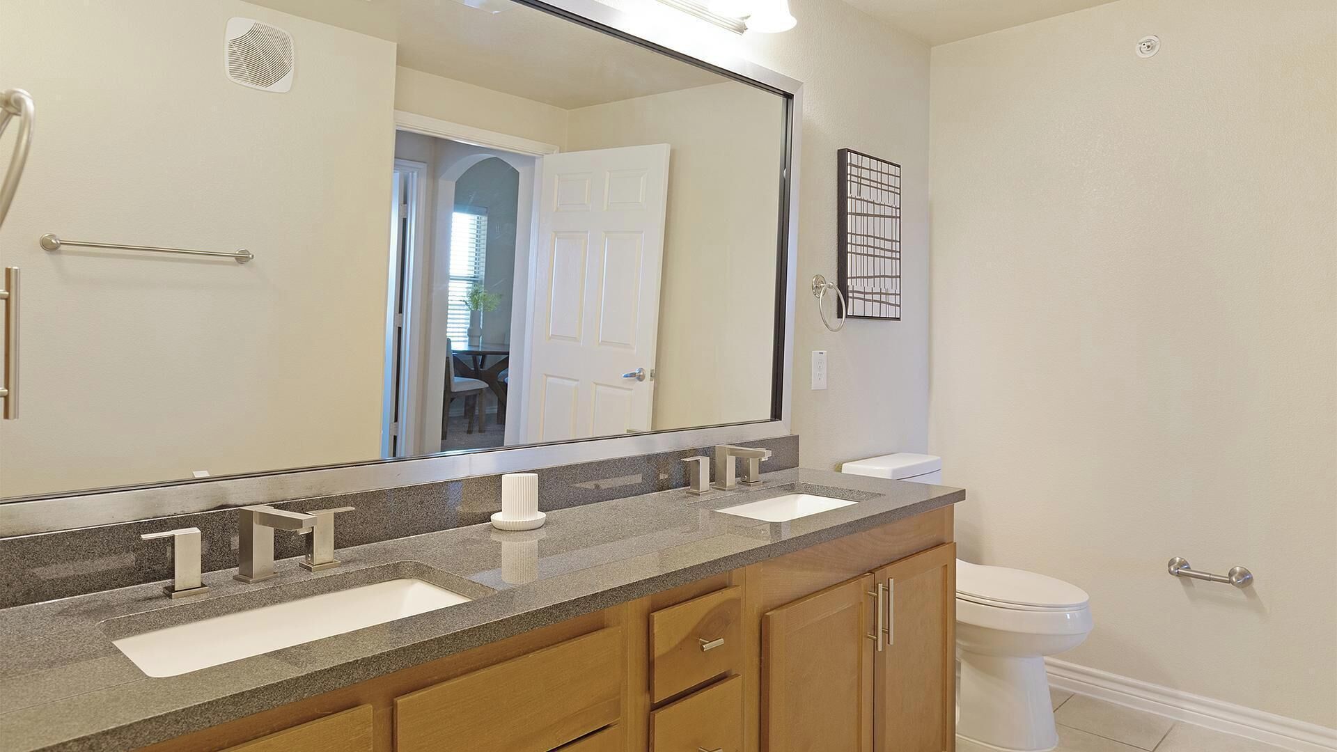A bathroom with a large mirror, two sinks, wooden cabinets, and a toilet.