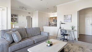 Uma sala de estar moderna com um sofá cinza, uma mesa de centro e uma escrivaninha com uma cadeira. Cozinha visível ao fundo, com armários e um micro-ondas.