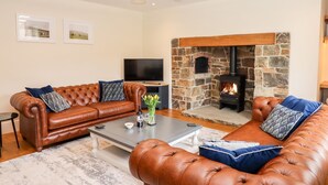 A cozy living room with a stone fireplace, leather sofas, a coffee table, and a flat-screen TV. - Knowle House (Okehampton)
