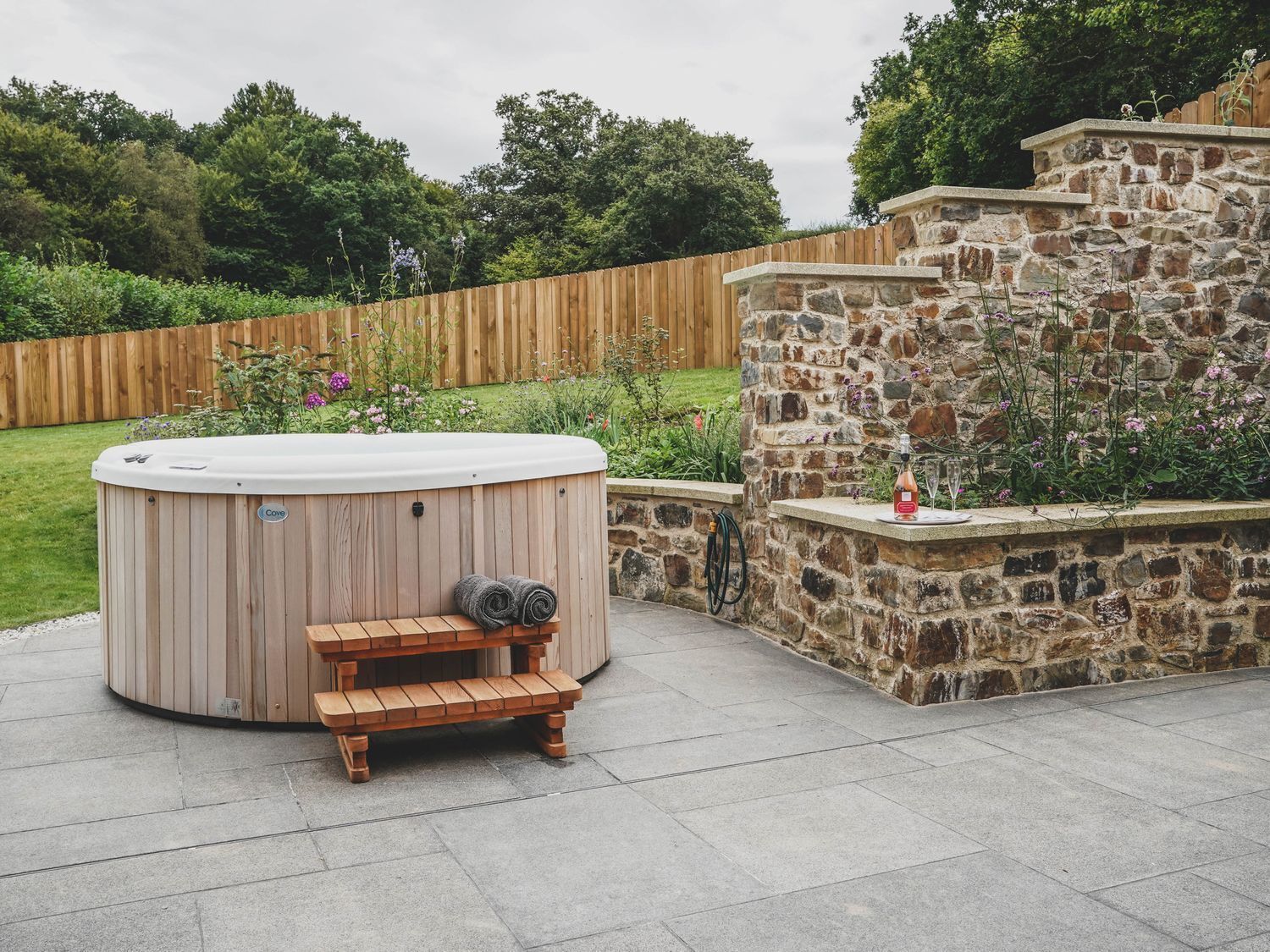 Een jacuzzi met een houten bank en een stenen muur met een tafeltje en bloemen.