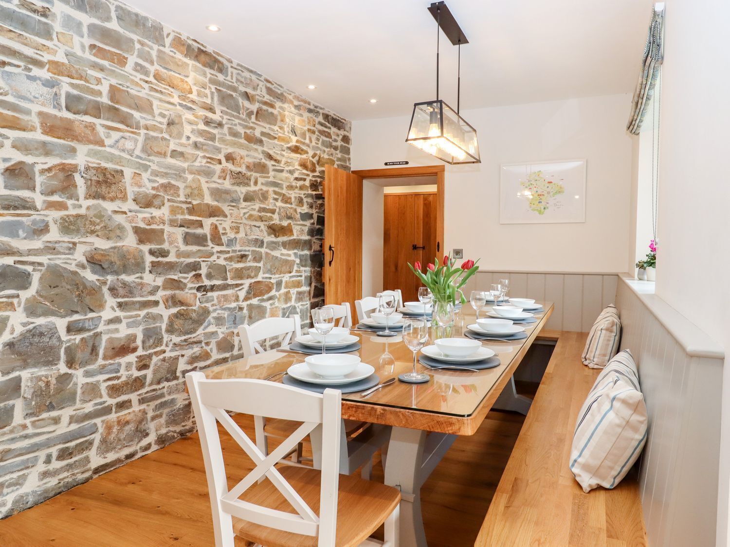 Un comedor con pared de piedra, mesa de madera, sillas blancas y una lámpara colgante.