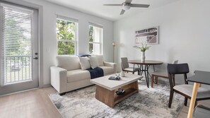 A modern living room with a sofa, coffee table, and chairs. There are large windows and a ceiling fan. - Landing | Beautiful Studio, Game Room, Gym (Jacksonville)