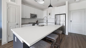 A modern kitchen with white cabinetry, a stainless steel refrigerator, a microwave, and a large island with bar stools. - Landing | Incredible 1BD, Gym, Clubhouse (Austin)