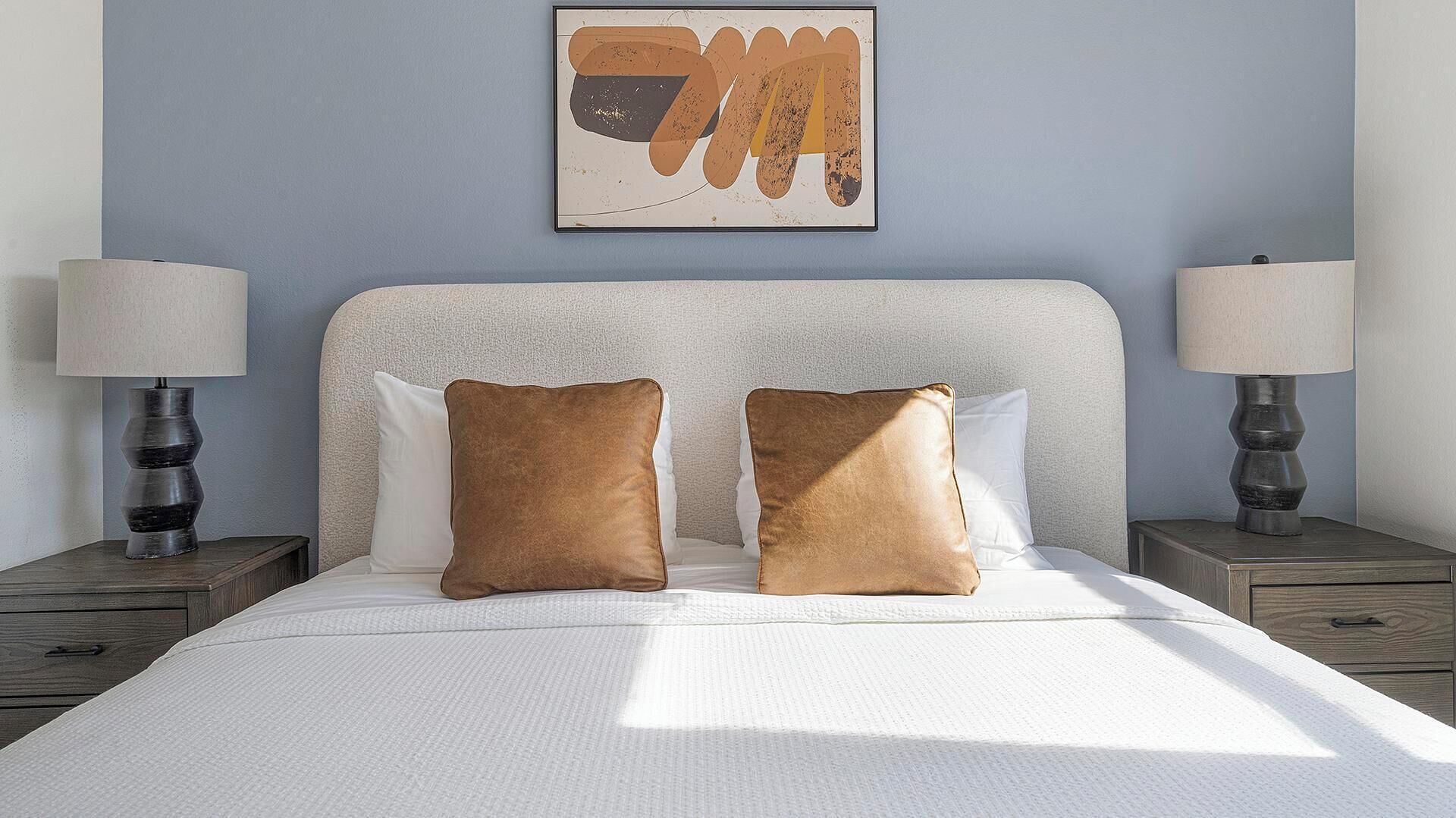 Een net opgemaakt bed met twee bruine kussens, een wit hoofdbord en twee bedlampen op houten nachtkastjes.
