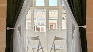 A balcony with two white chairs, green curtains, and a view of buildings through the windows. - Casa Maltija Sliema 4 Bedroom (Sliema)